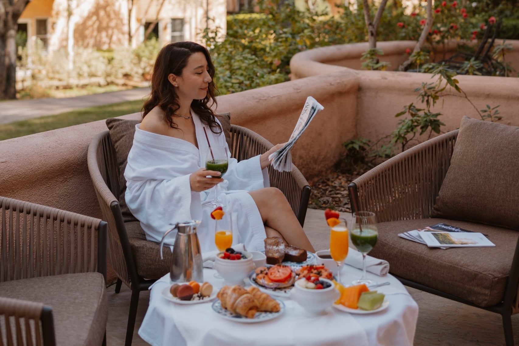 Woman sitting on a patio in a bathrobe reading a magazine next to a table full of breakfast food
